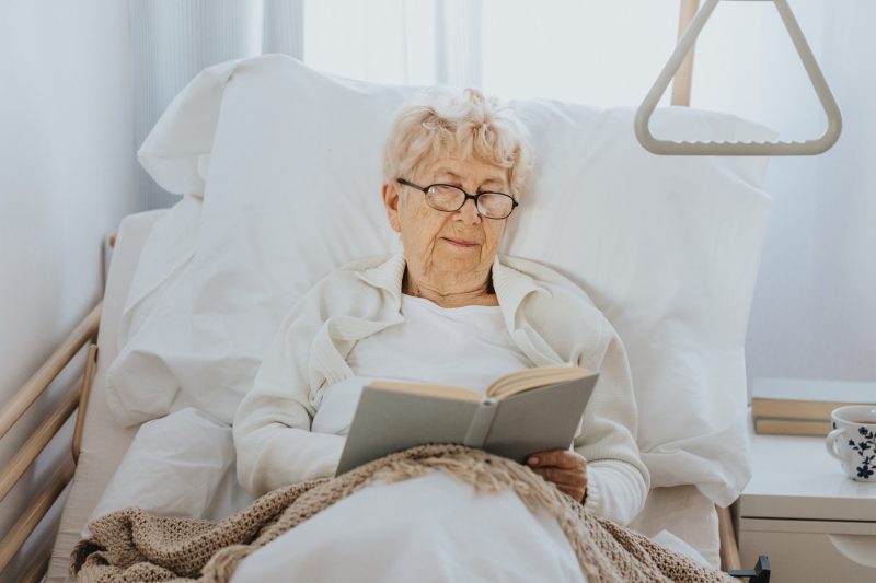 Seniorin liegt im Pflegebett und liest ein Buch