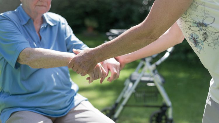 Mobilität in der Pflege | Älter Frau motiviert alten sitzenden Mann im Garten zum Aufstehen