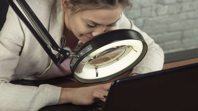 Optische Sehhilfen - Frau mit elektrisch beleuchteter Standlupe vor dem Laptop